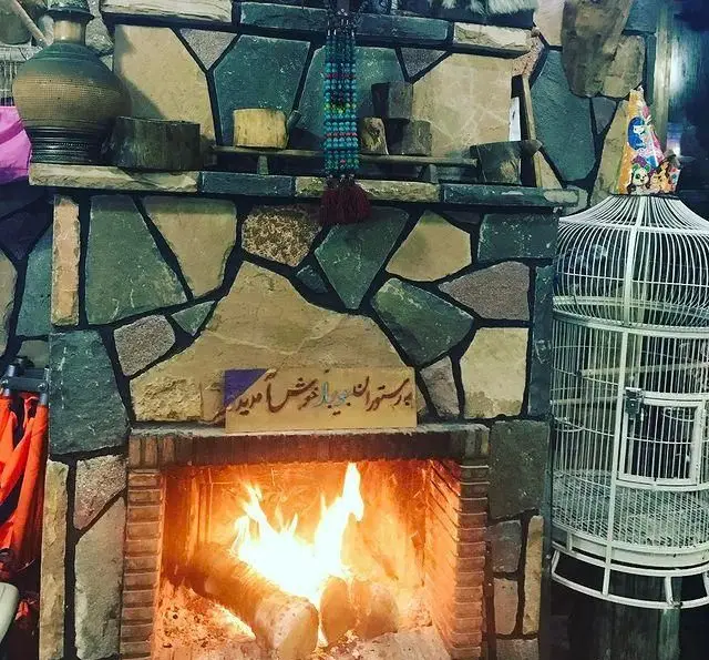 شومینه سنگی گرم و دنج در رستوران جویبار، با آتش فروزان، دیوارهای روستیک، دکوراسیون سنتی، قفس پرنده و خوش‌آمدگویی.