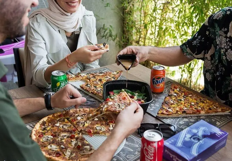 سه نفر در رستوران فست فود سیب ۳۶۰ مشغول خوردن پیتزاهای متنوع، سالاد تازه و نوشیدنی‌های خنک هستند و از دورهمی خود لذت می‌برند.