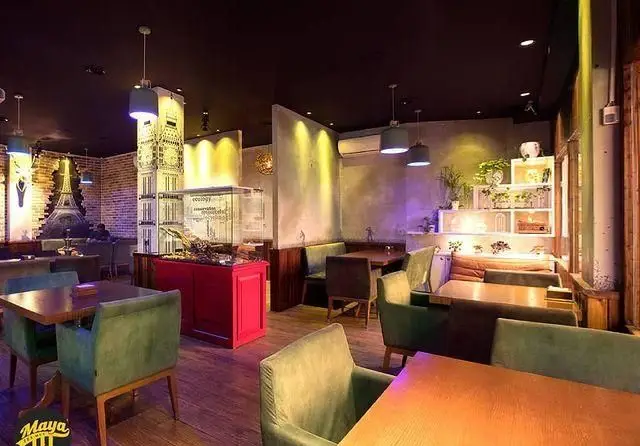 فضای داخلی دنج فست فود مایا تکس مکس با مبلمان راحتی سبز، دیوار آجری با طرح ایفل، برج ساعت و محفظه شیشه‌ای دکوراتیو.