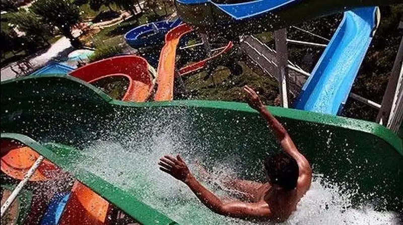 پارک آبی آزادگان: فردی در حال سر خوردن پرهیجان از سرسره آبی سبز رنگ با پاشش آب، نمایی از تفریحات هیجان‌انگیز تابستانی.