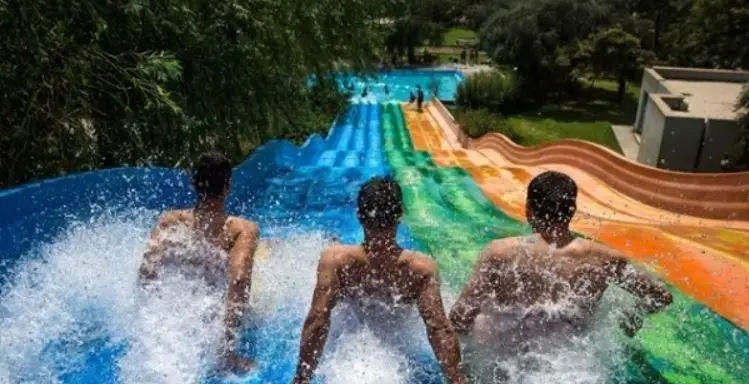 سه نفر در حال لذت بردن از هیجان سر خوردن از سرسره‌های آبی چند رنگ و بزرگ در پارک آبی آزادگان تهران، تابستانی شاد.