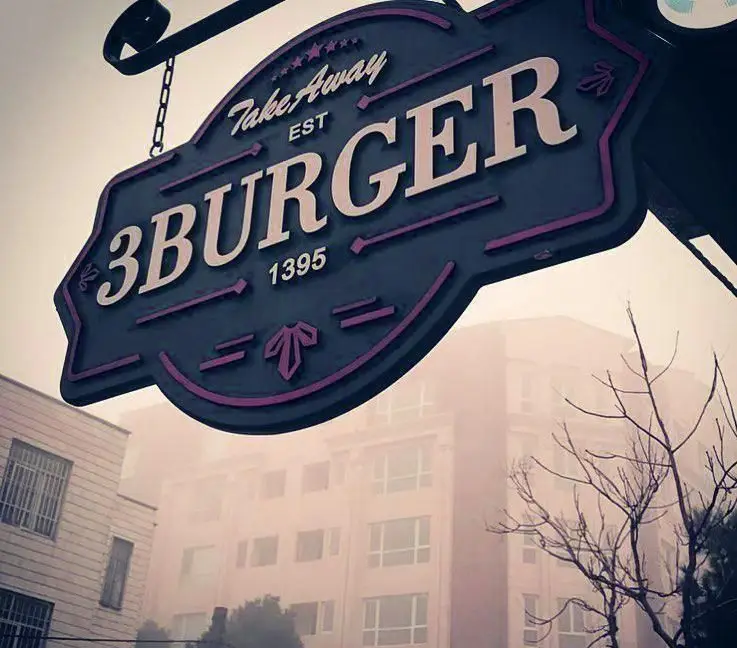 تابلوی آویز و برجسته "3BURGER" با عبارت "Take Away" و تاریخ "EST 1395" در یک روز مه آلود. این تصویر سردر رستوران فست فود و همبرگر را نشان می دهد.