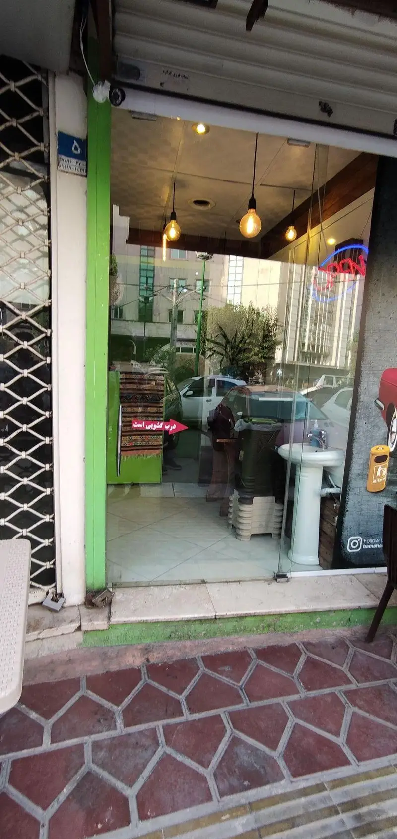 نمای بیرونی فست فود باماهاس با درب شیشه‌ای ورودی، چراغ‌های آویز و انعکاس خیابان. تصویری جذاب از ظاهر مغازه.