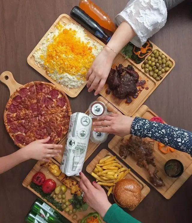 دورهمی شلوغ در فست فود باماهاس با تنوع غذایی بالا شامل پیتزا، برگر، کباب، سیب‌زمینی سرخ‌کرده و برنج.