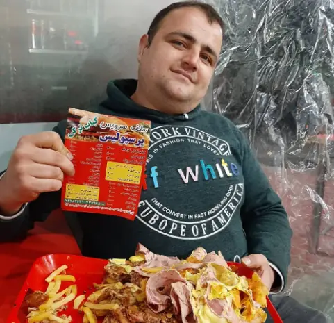 مردی در فست فود پرسپولیس منوی کباب ترکی را نشان می‌دهد، کنار سینی بزرگی از غذا شامل سیب‌زمینی سرخ کرده و ساندویچ‌های خوشمزه.