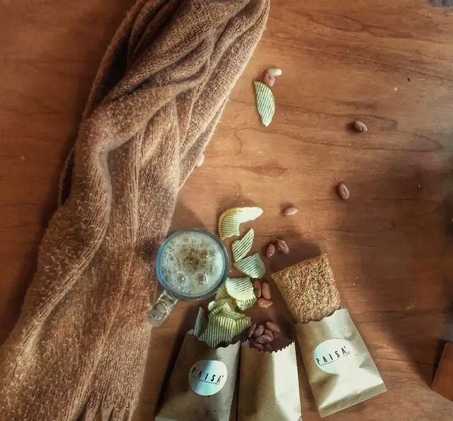 میز چوبی کافه پاییزا با نوشیدنی گرم، چیپس و آجیل در بسته‌بندی‌های اختصاصی PAISA، به همراه یک بار غلات برای میان‌وعده سالم.