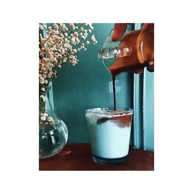 آماده سازی نوشیدنی لاته یا موکا در کافه پاییزا؛ دستی در حال ریختن قهوه روی شیر در لیوانی کنار گلدان گل‌های صورتی. کافه پاییزا