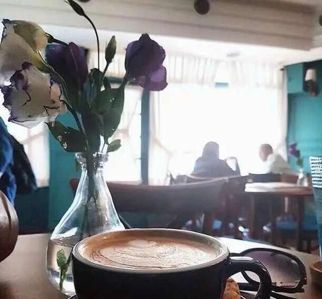 نمایی دنج و دلنشین از کافه پاییزا با یک فنجان لاته داغ و طرح‌دار، گلدان گل‌های تازه و مشتریانی در حال گپ و گفتگو. ایده‌آل برای آرامش.