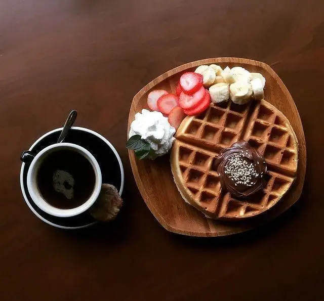 صبحانه دلپذیر کافه پاییزا: وافل طلایی با توت فرنگی، موز، خامه، شکلات و کنجد، در کنار یک فنجان قهوه تازه دم.
