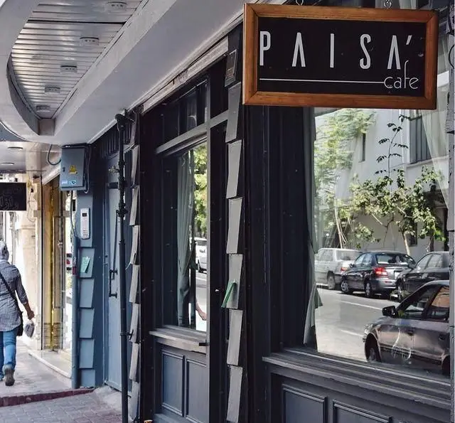 نمای بیرونی کافه پاییزا با تابلوی مشکی و لوگوی سفید، انعکاس خیابان و ماشین‌ها در شیشه، محیطی دنج و آرام برای مشتریان.