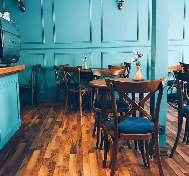 دکوراسیون داخلی دنج و شیک کافه پاییزا با دیوارهای آبی فیروزه‌ای، کفپوش چوبی و صندلی‌های چوبی با کوسن‌های آبی مخملی. محیطی ایده‌آل برای استراحت.