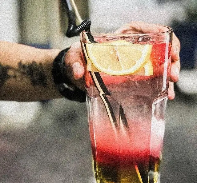 نوشیدنی لایه‌ای و رنگارنگ کافه پاییزا شامل شربت قرمز و لایه شفاف، با تزیین لیمو، توت فرنگی و نی مشکی، طعمی خنک و دلنشین.