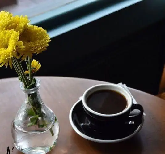 یک فنجان قهوه مشکی روی نعلبکی همراه با گلدان کوچک گل‌های زرد روی میز چوبی. لحظات دلنشین در کافه پاییزا، انتخابی ایده‌آل برای استراحت.