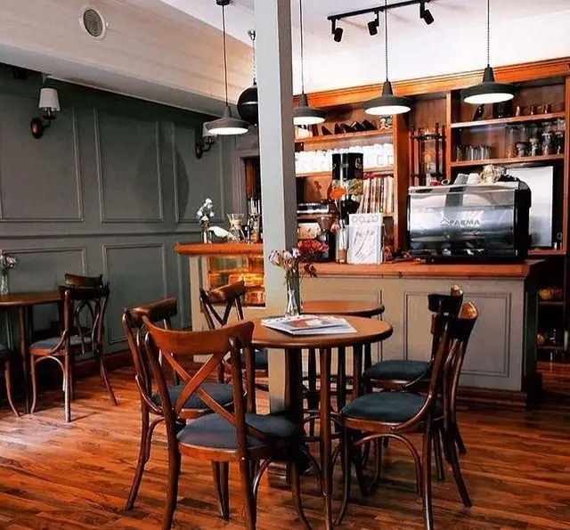 کافه پاییزا: دکوراسیون داخلی دلنشین با میزهای چوبی، صندلی‌های راحت، دستگاه قهوه FARMA، پیشخوان مرتب و قفسه‌های تزئینی پر از جزئیات.
