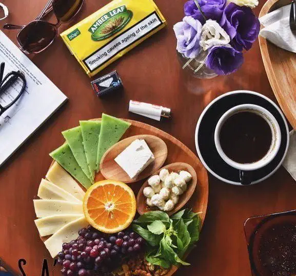 میز صبحانه/عصرانه در کافه پاییزا: قهوه، پنیرهای متنوع، میوه‌های تازه (انگور، پرتقال) و چیدمان گل. فضایی آرامبخش.