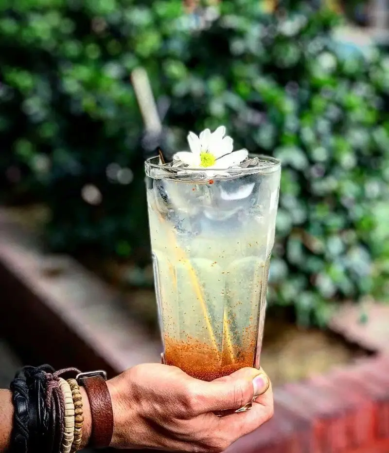 نوشیدنی خنک و دلچسب کافه پاییزا با گل سفید روی لیوان، در دست با دستبندهای چرمی و چوبی. حس تازگی در فضای سبز کافه.