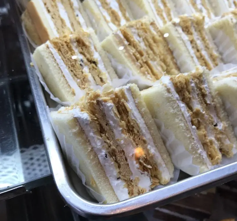 نمای نزدیک از سینی ساندویچ‌های شیرین و خوشمزه کافه قنادی لادن با لایه‌های خامه و مغز ترد، آماده سرو در ویترین.