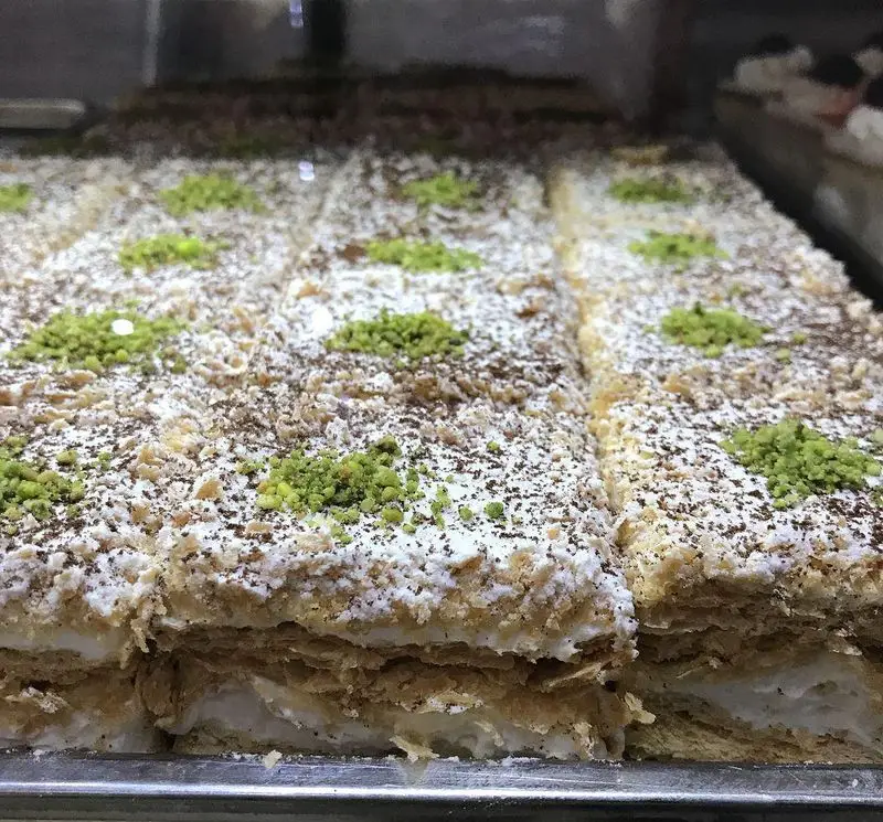 دسر لذیذ و تازه کافه قنادی لادن: شیرینی چندلایه با کرم لطیف، تزئین شده با پودر قند، خرده بیسکوییت و پودر پسته.