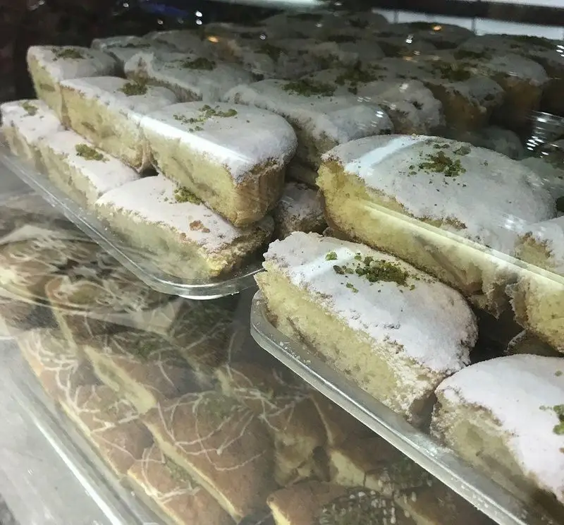 ویترین جذاب و متنوع کافه قنادی لادن: شیرینی‌های تازه و کیک‌های خوشمزه با پودر قند و پسته، مناسب برای سفارش شما.