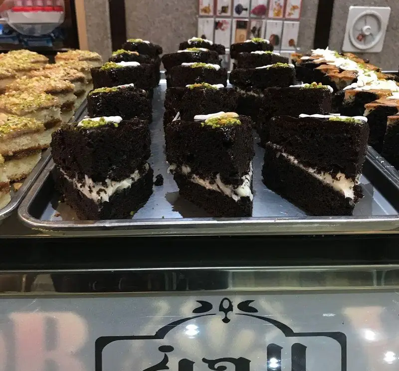 تصویری از سینی‌های پر از کیک شکلاتی تازه با لایه کرم سفید و تزئین پسته، و شیرینی‌های سنتی در ویترین کافه قنادی لادن.