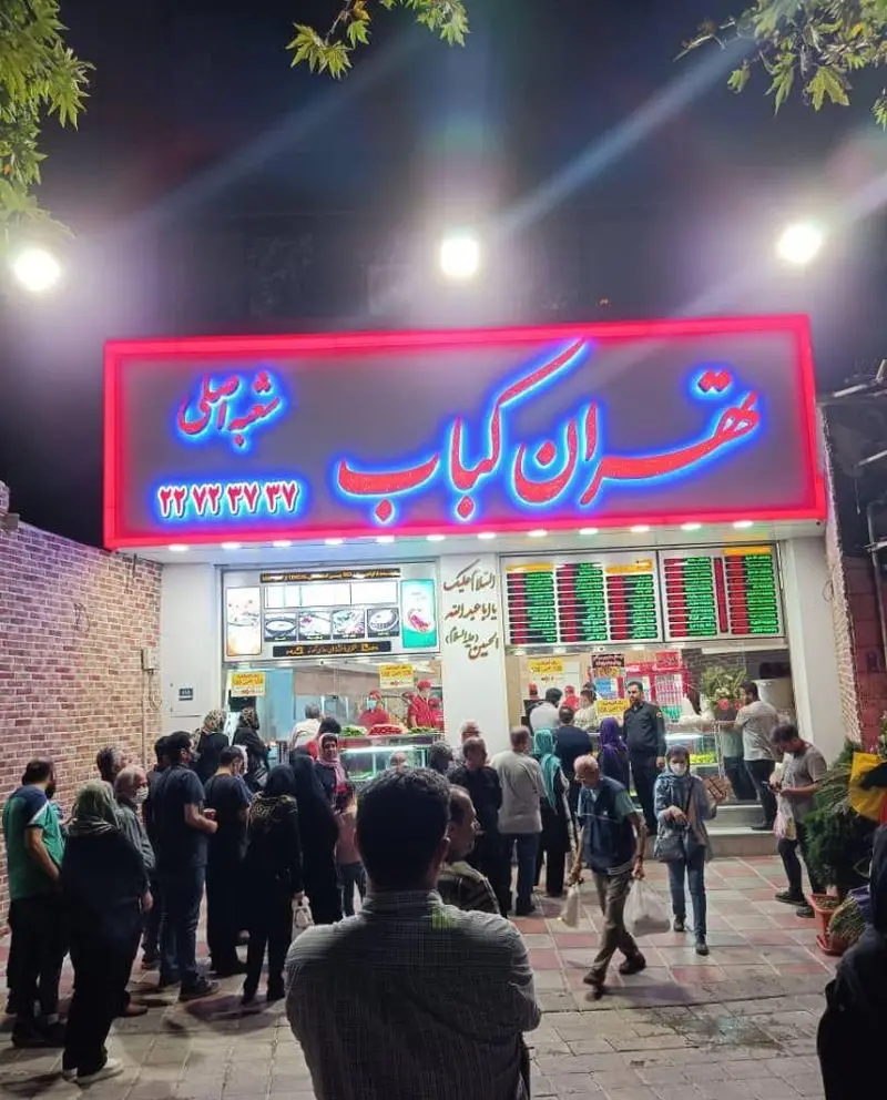 تصویر رستوران شلوغ تهران کباب در شب، با تابلوی نئون درخشان "تهران کباب" و شماره تماس، صف مشتریان برای خرید.