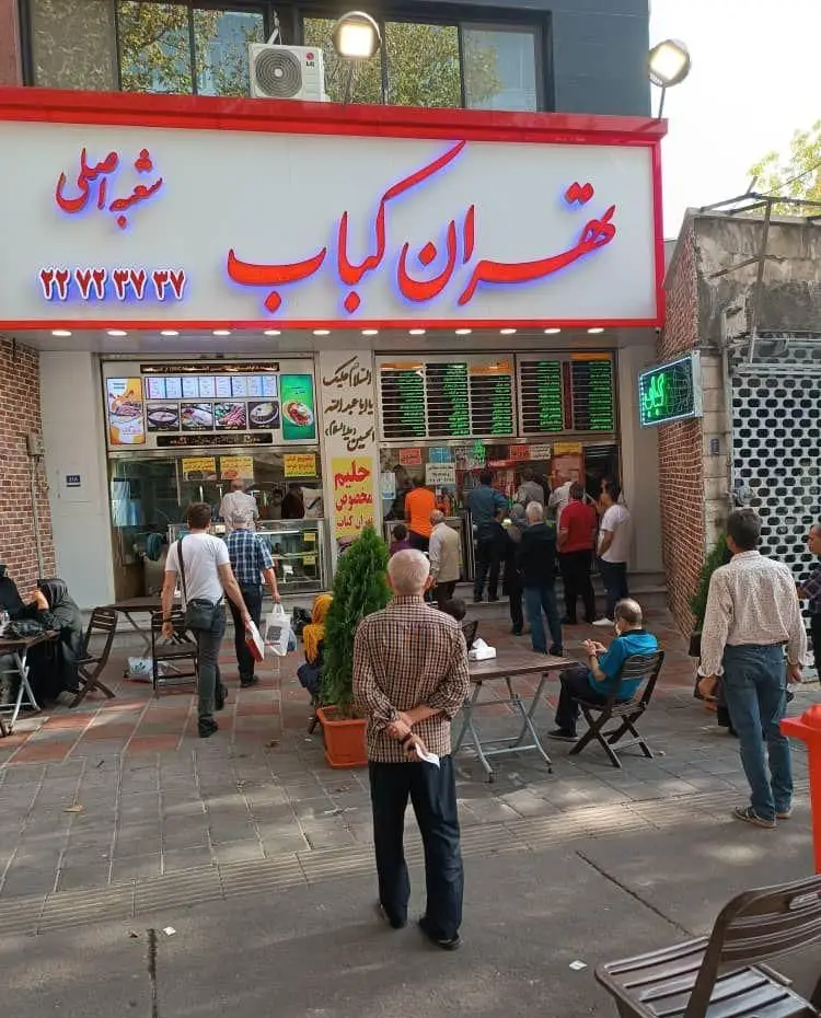 تصویری از نمای بیرونی رستوران شلوغ تهران کباب، شعبه اصلی با تابلوی نئون قرمز و آبی، مشتریان در صف و صندلی‌های بیرون.