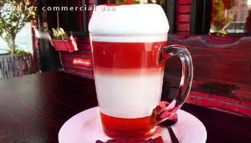 تصویری زیبا از نوشیدنی لایه‌ای با رنگ‌های جذاب قرمز و سفید و خامه فراوان، سرو شده در لیوان شیشه‌ای دسته دار در فضای دلنشین کافی شاپ چهارچوب.