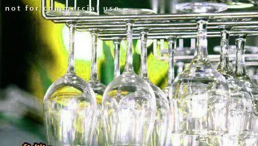 نمایی نزدیک از لیوان های گیلاس تمیز و شفاف که به صورت وارونه از قفسه فلزی در محیط دنج کافی شاپ چهارچوب آویزان شده‌اند.