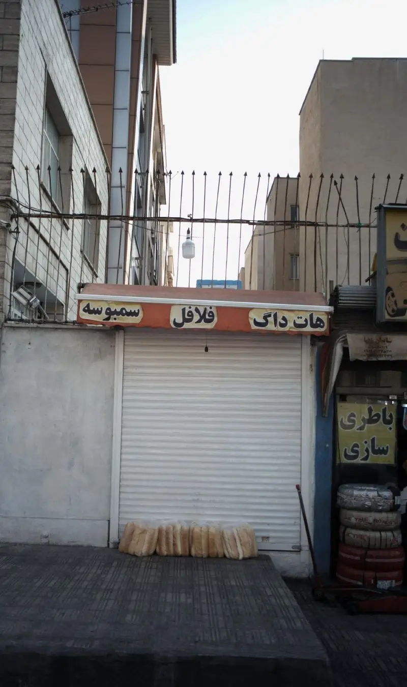 تصویری از فروشگاه فیت برگر (Fit Burger) که ساندویچ‌های هات داگ، فلافل و سمبوسه ارائه می‌دهد. کرکره مغازه پایین و نان‌ها بیرون هستند.