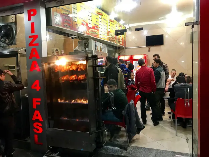 نمای بیرونی رستوران پیتزا چهار فصل. تابلوی قرمز "PIZZA 4 FASL"، دستگاه جوجه گردان با مرغ‌های بریان و مشتریان مشغول غذا.
