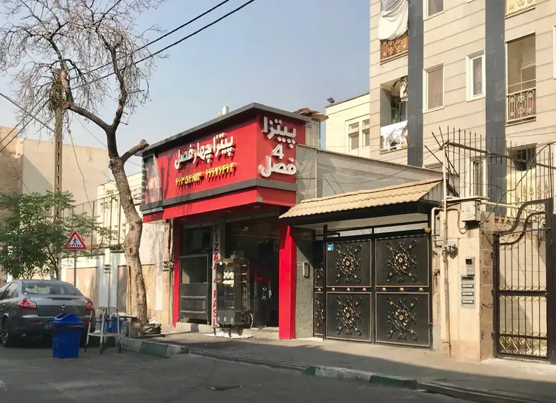 نمای بیرونی رستوران پیتزا چهار فصل در یک روز آفتابی با تابلوی قرمز رنگ و خودروی پارک شده در خیابان