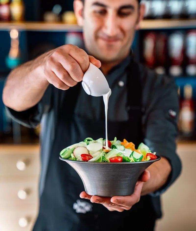 سرآشپز اینجا برگر در حال سرو یک سالاد تازه و سالم با سس مخصوص، نشان‌دهنده تنوع و کیفیت غذا در رستوران ما.