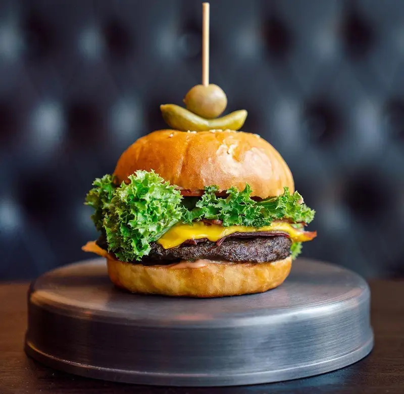 تصویری از برگر ویژه اینجا برگر، با نان تازه، گوشت لذیذ، پنیر چدار آب شده، کاهو و بیکن، تزئین شده با زیتون و خیارشور روی پایه فلزی.