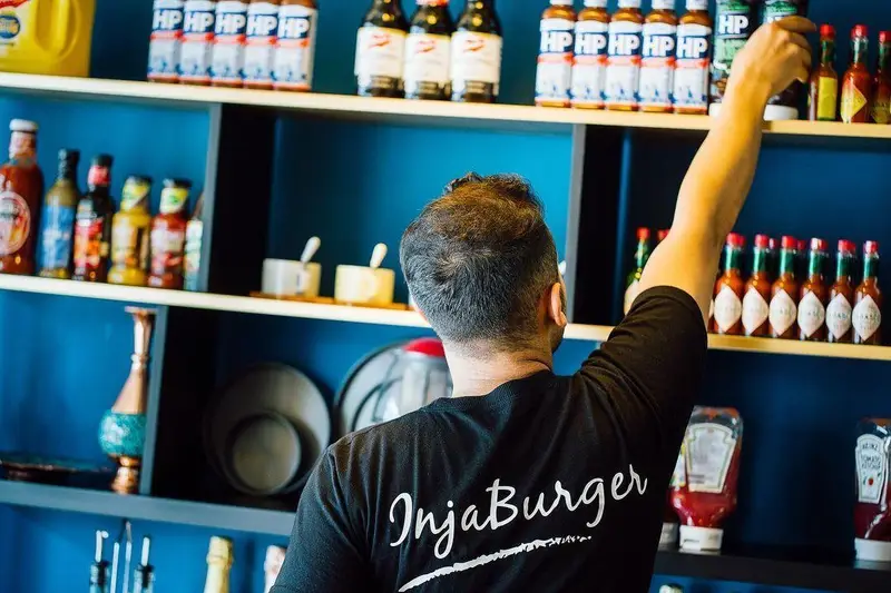 تصویری از کارمند رستوران اینجابگر که در حال برداشتن بطری سس از قفسه است. نمایش تنوع سس‌ها و مواد اولیه باکیفیت اینجا برگر.