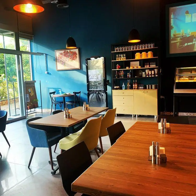 تصویری از فضای داخلی رستوران اینجا برگر با دیوار آبی، میزهای چوبی، صندلی‌های مدرن، قفسه‌های دکوری و نورپردازی دلنشین.