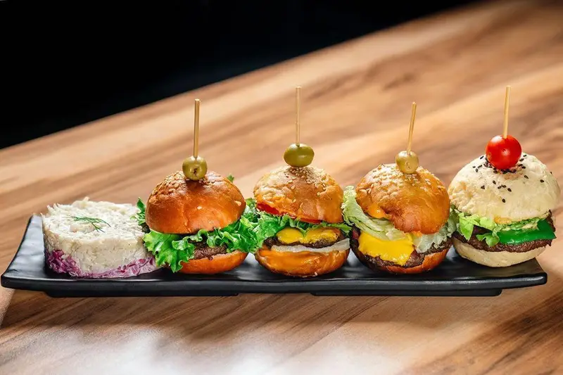 سینی مزه بی‌نظیر اینجا برگر شامل چهار مینی همبرگر اسلایدر خوشمزه و متنوع با نان‌ها و مواد تازه، همراه با سالاد جذاب.