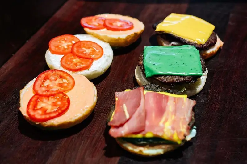 آماده‌سازی مینی برگرهای متنوع اینجا برگر با نان تازه، گوجه، سس‌های مخصوص، پنیرهای رنگی زرد و سبز و تاپینگ‌های جذاب