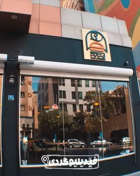 تصویری از نمای بیرونی فست فود اینجا برگر، یک رستوران مدرن با تابلوی لوگوی جذاب و پنجره‌های بزرگ شیشه‌ای که محیط شهری را بازتاب می‌دهند.