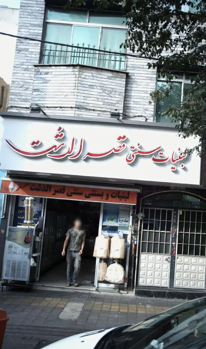 نمای خارجی مغازه لبنیات و بستنی سنتی قصرالدشت با تابلو روشن و محصولات محلی، در یک خیابان شلوغ شهری در ایران.