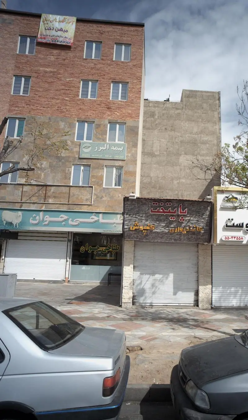 عکس از نمای بیرونی طباخ جوان، رستوران یا کبابی در خیابان، با تابلوی آبی و لوگوی گوسفند و کرکره سفید بسته.