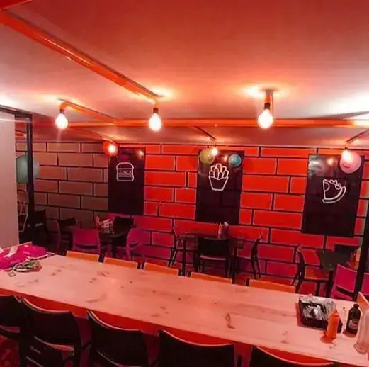 فضای داخلی مدرن و جذاب فست فود بشکه با دیوارهای آجری قرمز، نورپردازی گرم، میزهای چوبی بلند و نمادهای خوشمزه برگر و سیب زمینی.