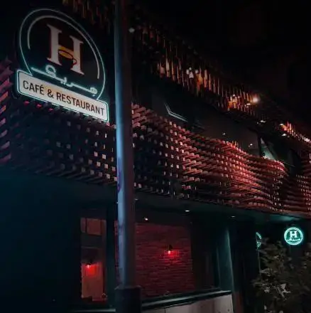 تصویر نمای بیرونی کافه رستوران هریک در شب با تابلوی نئونی بزرگ “H” و فنجان قهوه، همراه با نوشته کافه و رستوران. طراحی مدرن و چشم‌نواز.