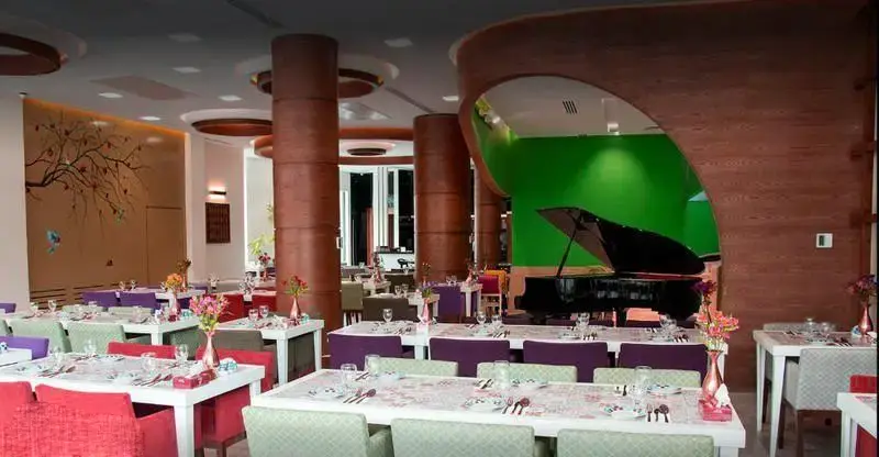 نمای داخلی کافه رستوران گارنیش با میز و صندلی‌های چیدمان شده، پیانو گرند مشکی در بخش سبز رنگ و نقاشی درخت و پرنده روی دیوار.