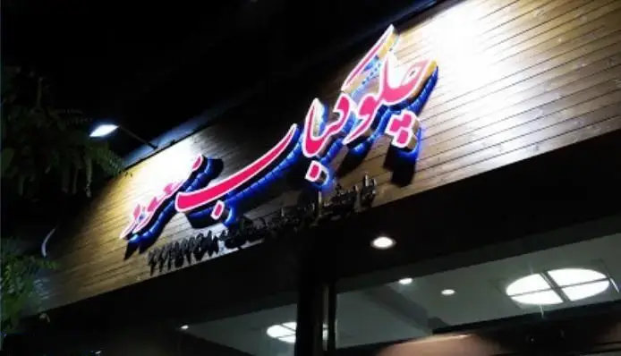 نمای شبانه و روشن از تابلوی نئون رستوران مسعود با نورپردازی صورتی و آبی روی دیوار چوبی. ورودی رستوران با بازتاب نور داخلی.