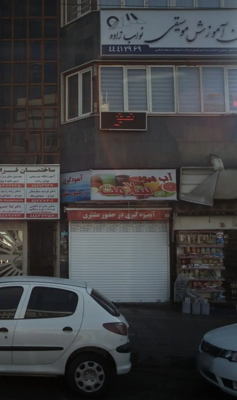 نمای خیابانی از آبمیوه فروشی سلامت با تابلو 'آبمیوه گیری در حضور مشتری'. بالای آن آموزشگاه موسیقی نواب زاده و سایر کسب و کارها دیده میشود.
