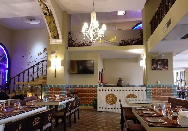 فضای داخلی رستوران سنتی عمارت فردوس با دکوراسیون ایرانی-اسلامی، شامل پلکان، خوشنویسی، دیوار آجری و میزهای آماده پذیرایی با ظروف مسی.