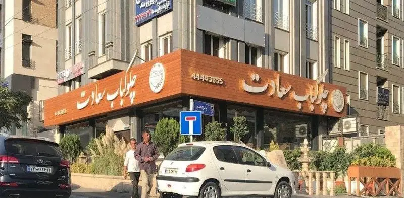 تصویری از نمای بیرونی رستوران چلاب سعادت، با تابلوی چوبی و شماره تماس 44443855، در خیابانی شلوغ در شهر تهران.
