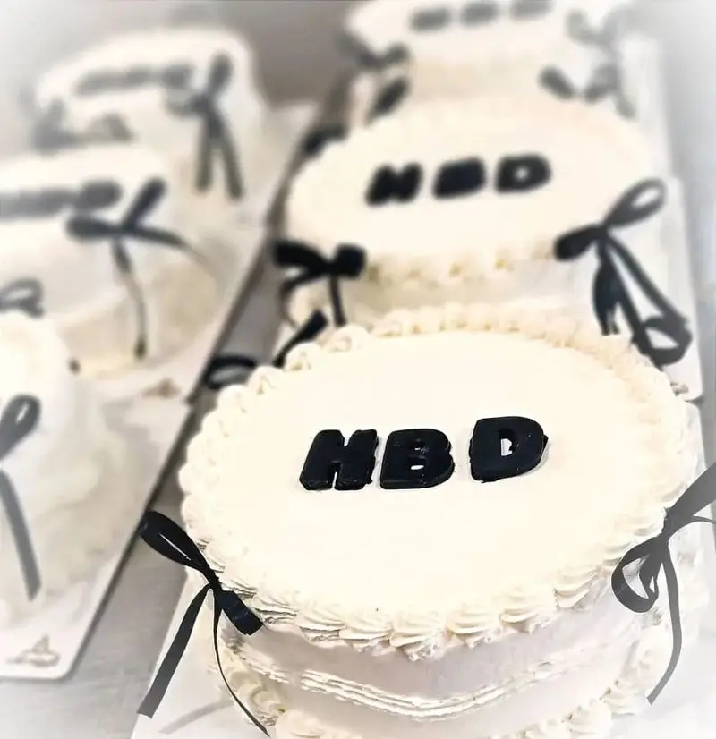 نمایی از چندین کیک تولد سفید با تزیین روبان مشکی و متن HBD، آماده در قنادی شیرینی الف شعبه تهرانپارس.