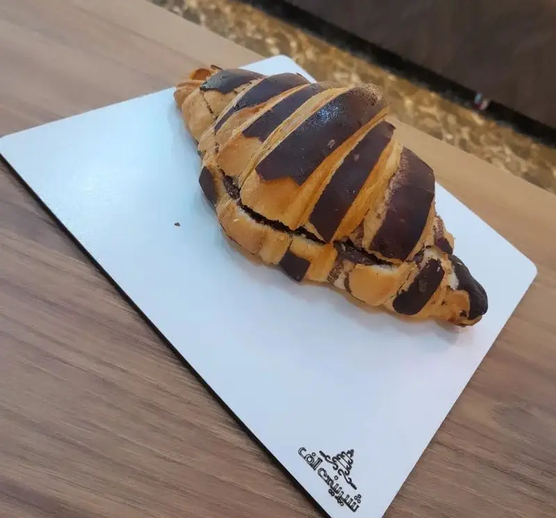 کروسان شکلاتی لذیذ و تازه با تزئینات کاکائویی روی تخته سفید، محصول انحصاری شیرینی الف شعبه تهرانپارس.