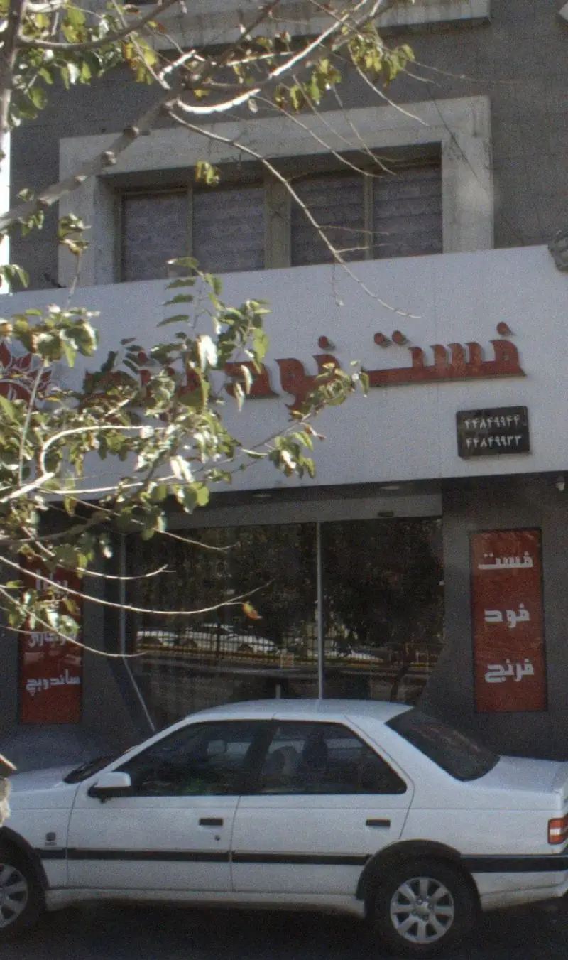 نمای بیرونی فست فود فرنج با تابلوی قرمز رنگ، شماره تلفن و یک خودروی سفید پارک شده در مقابل ورودی، همراه با درختان.